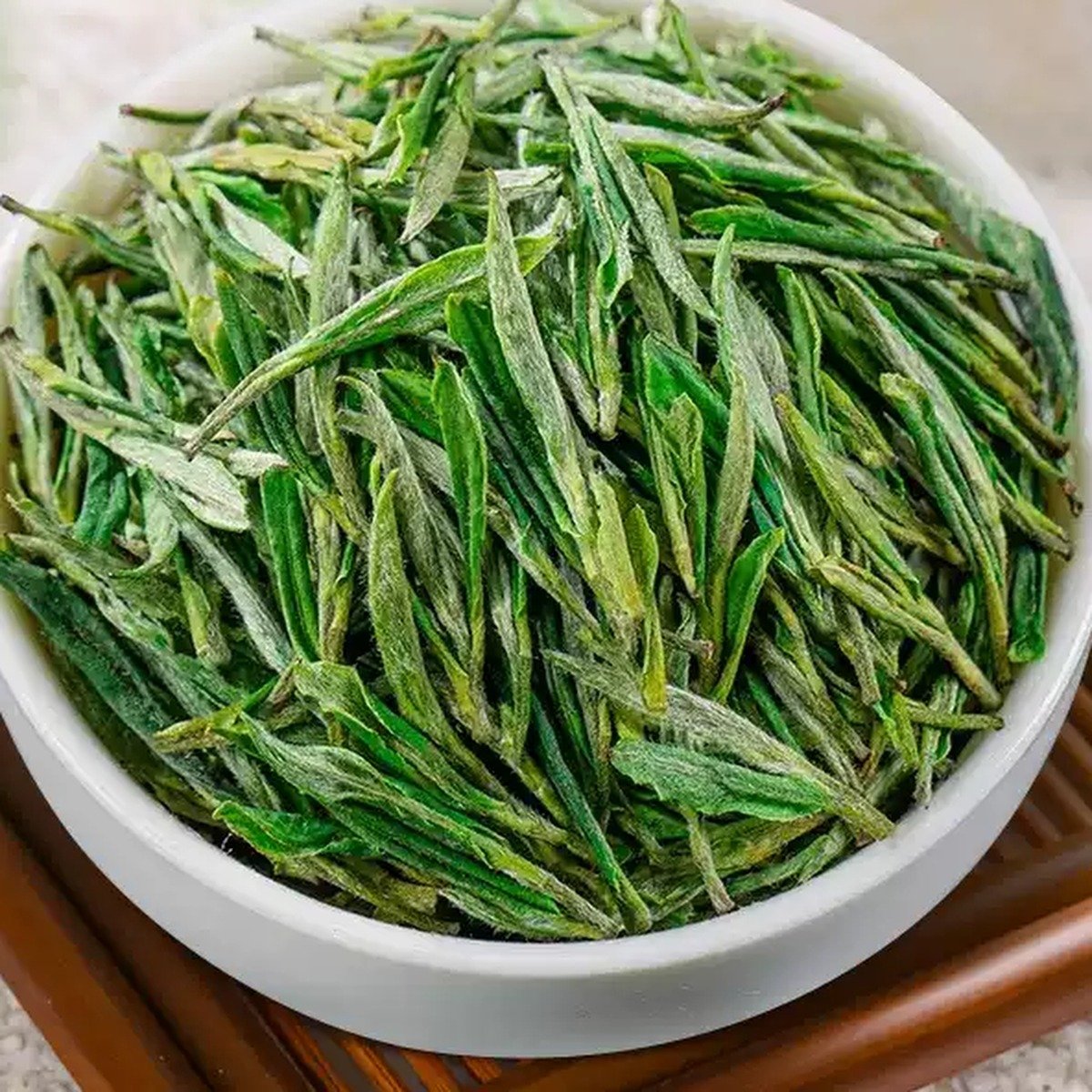 White bowl filled with slender downy Huangshan Maofeng dry green tea leaves on a wooden slatted tray