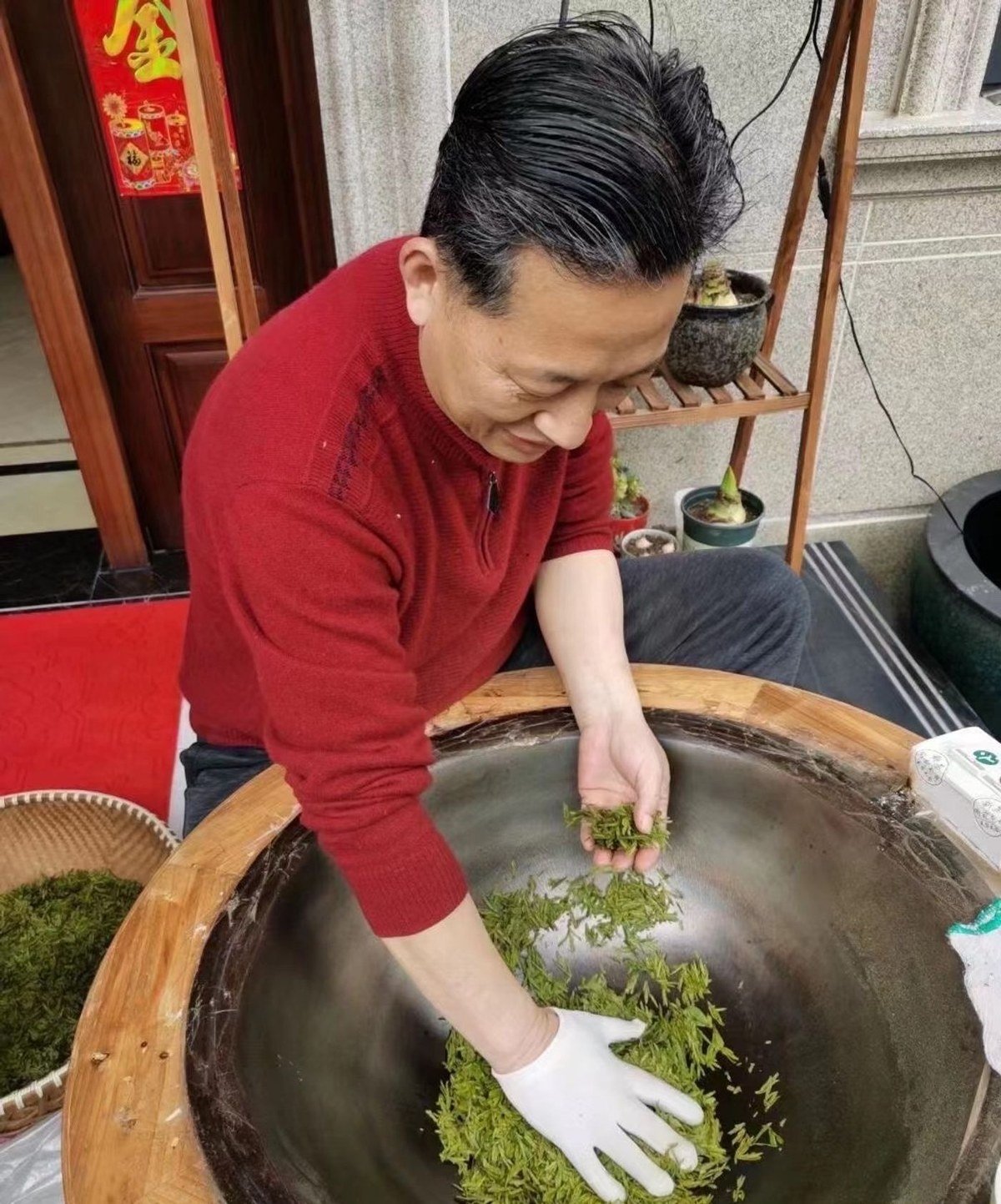 A tea maker turning bright green leaves by hand in a traditional iron wok during wok-firing, with a basket of fresh leaf beside the pan